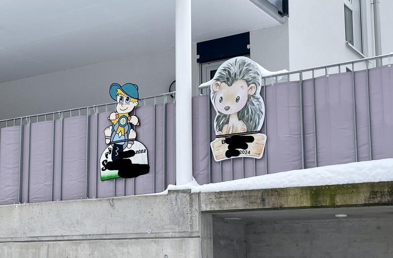Twee babygeboortebordjes opgehangen aan een grijs balkon. De namen en precieze geboortedatum zijn geanonimiseerd, alleen de eerste letter van de naam en het geboortejaar zijn zichtbaar. Het linker bord is van een kindje S. dat in 2022 is geboren. Het stripfiguur op het bord is van een blond jongetje met een groene pet op een scooter. Het rechter bord is van B. die in 2024 is geboren. Boven de naam staat een grote cartooneske egel.