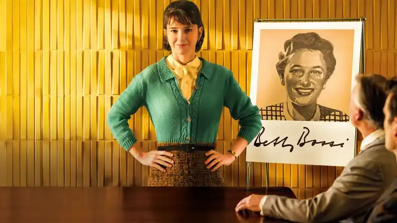 Actrice Sarah Spale geeft een presentatie voor een geel betegelde muur en naast een flipover met een A1 grote zwart-wit afbeelding van het Betty Bossi karakter. Ze staat met haar armen in de zij voor een tafel waar twee mannen in pak aanzitten.