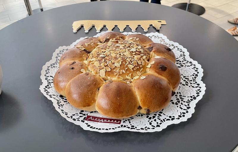 Een grijze tafel met daarop een bruin glanzend driekoningenbrood. Op het brood zit amandelschaafsel en er steken wat stukjes chocolade door de korst. Achter het brood ligt een gouden kroon in nog in opgevouwen toestand.
