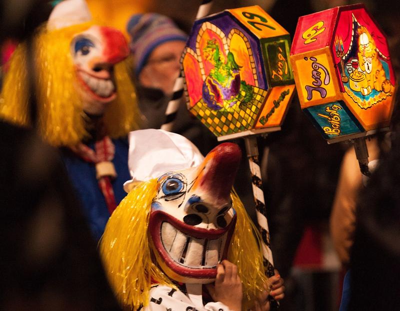 Foto van een kind met een traditioneel masker en een lampion tijdens de Basler Fastnacht. Het masker is een karikatuur met grote ogen, neus en mond en bedekt het gehele gezicht. Op de achtergrond is nog iemand met een masker te zien.