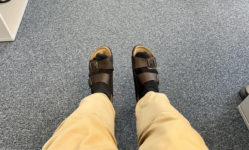 Van boven gefotografeerde onderbenen en voeten met twee bruine slippers die boven de grond zweven. De slippers zijn van kurk en hebben twee leren riemen met goudkleurige gespen. De broek is lichtbruin. De vloer is een blauwgrijs kantoortapijt.