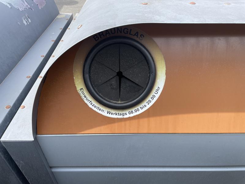 Een metalen container met een ronde opening, omgeven door een Duitse tekst die vertaald naar Inwerptijden: Werkdagen 08:00 tot 20:00 uur.
