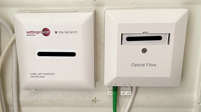 Twee witte vierkante glasvezelkastjes op een witte achtergrond. De linker heeft een sticker van wettingen4net en een waarschuwing