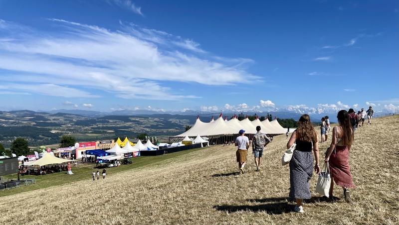 Mensen lopen over een heuvel naar een festivalterrein met witte tenten en kleurrijke kraampjes, tegen een achtergrond van een uitgestrekt landschap en blauwe lucht.