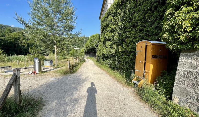 Een zonnig pad met schaduw van een persoon, omgeven door groene bomen en struiken. Rechts staat een houten wc-hokje tegen een met klimop begroeide muur.