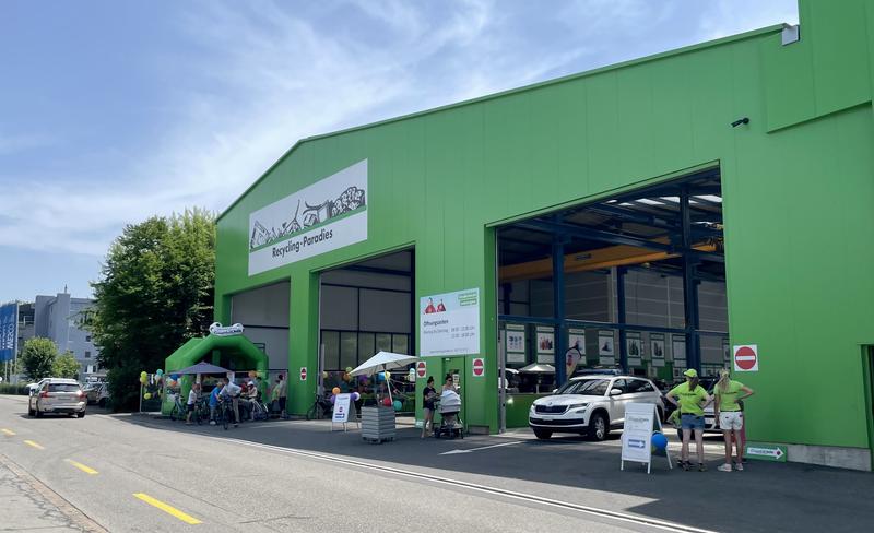 Een groen gebouw met een grote openstaande deur, waar auto's naar binnen rijden. Mensen staan buiten onder een groene tent. Boven de deur hangt een bord met de tekst Recycling Center.