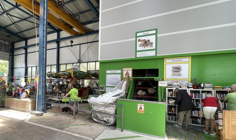 Een recyclingcentrum met mensen die boeken en andere items sorteren. Er zijn groene containers en planken met boeken.