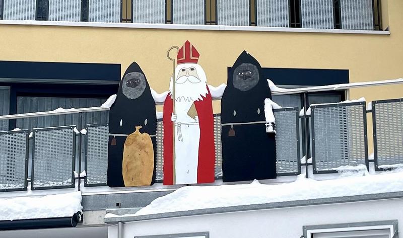 Drie borden met in het midden een cartoonesk getekende Samichlaus met rood gewaad, mijter en een staf. Aan zijn zijde twee Schmutzli's in zwarte gewaden, grijze baarden en een donker gezicht. De linker Schmutzli heeft een jute zak voor zich staan. De borden hangen aan de railing van een balkon met her en der een hoopje sneeuw op de randen van het balkon.