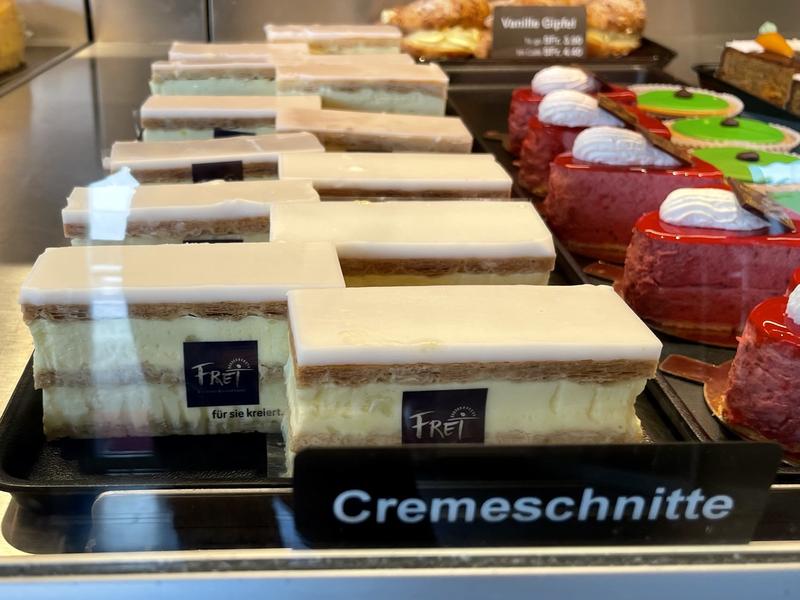 Een rij cremeschnitten achter een glazen vitrine. De gebakjes bestaan uit twee lagen crème en drie lagen bladerdeeg. Bovenop zit een dikke laag wit glazuur. De gebakjes liggen links en rechts verschoven achter elkaar.