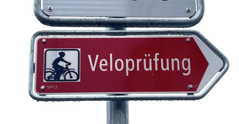 Rood verkeersbord met de tekst 'Veloprüfung' en een pictogram van een fietser, wijzend naar rechts.